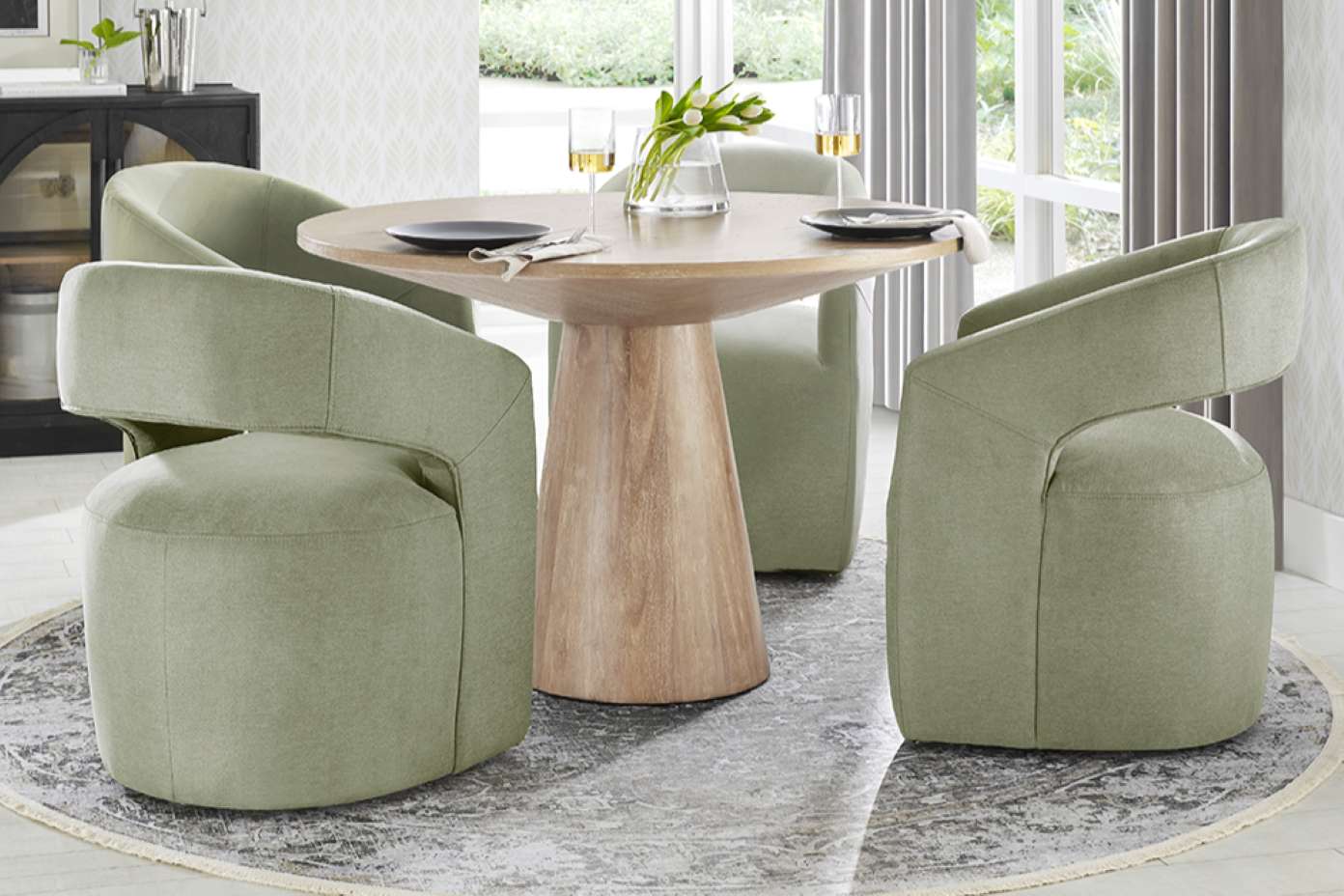 A round wooden dining table with four green upholstered chairs on a patterned rug.
