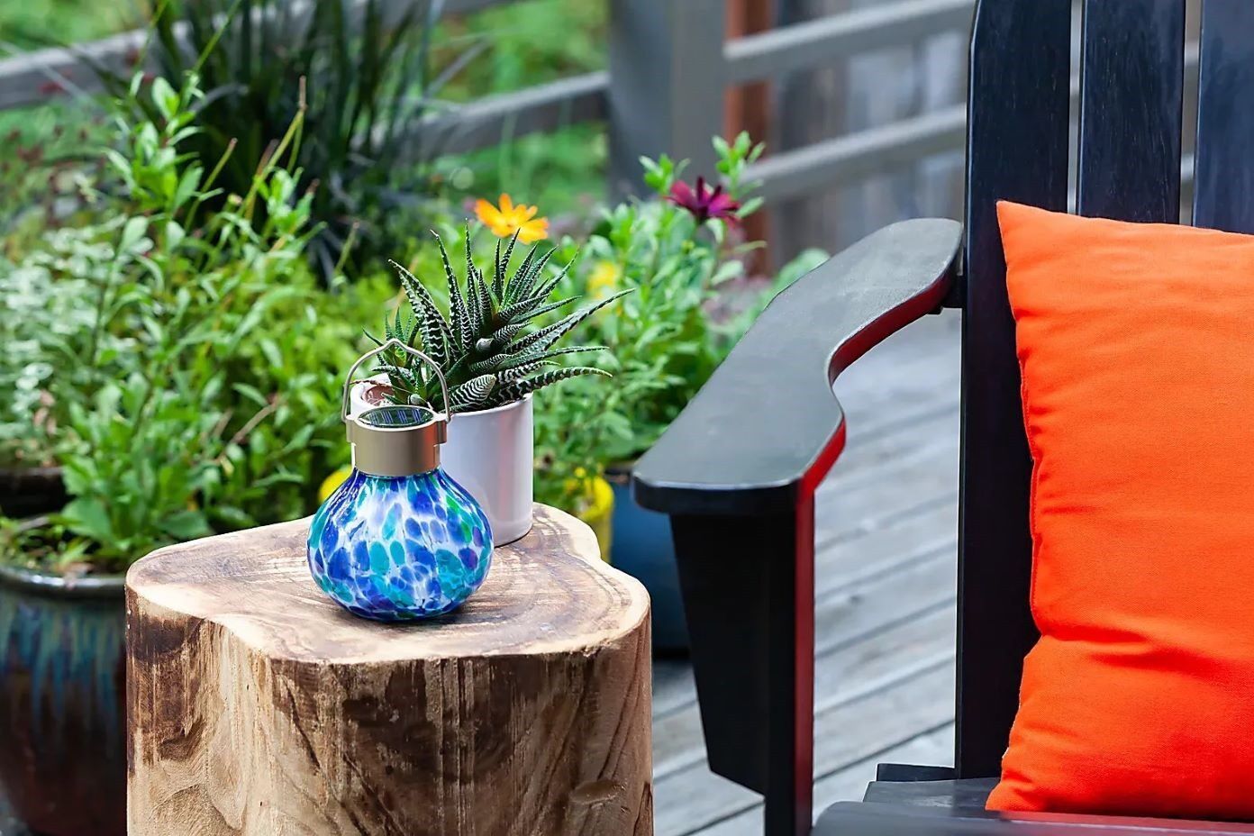 A blue and green glass lantern sits on a wooden stump next to a black chair with an orange pillow.
