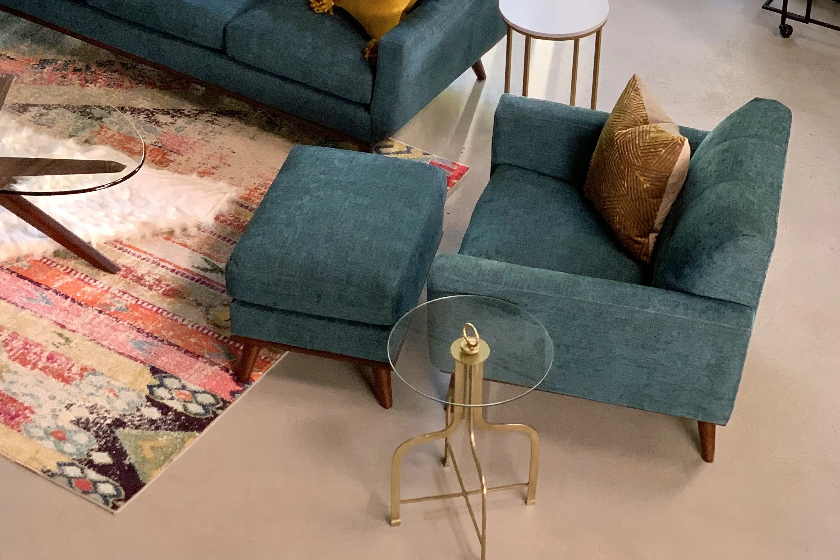 A teal armchair with a matching ottoman, a glass-topped side table, and a colorful patterned rug in a modern living room.
