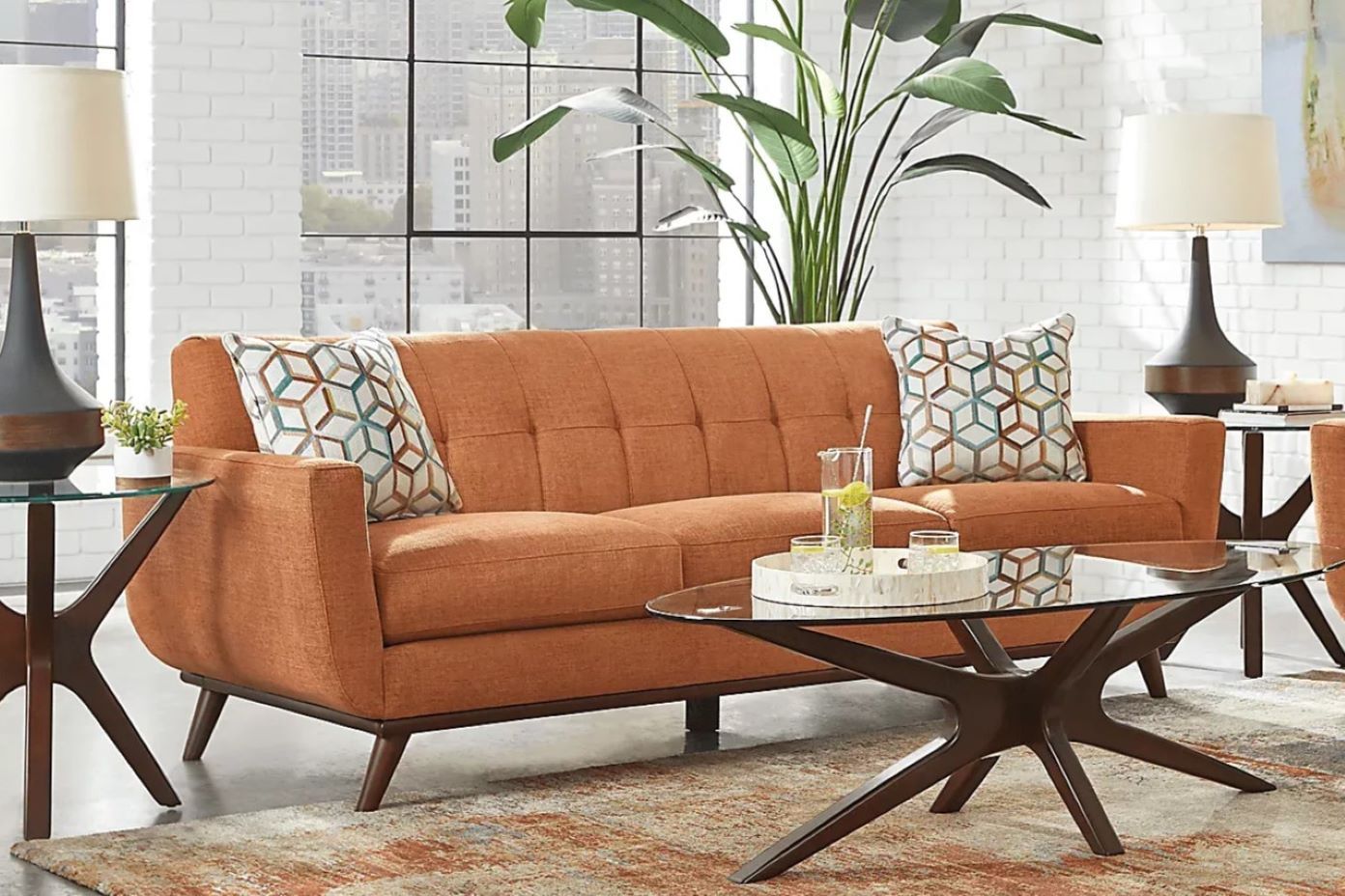 Orange mid-century modern sofa with geometric pillows, glass coffee table, and lamps.
