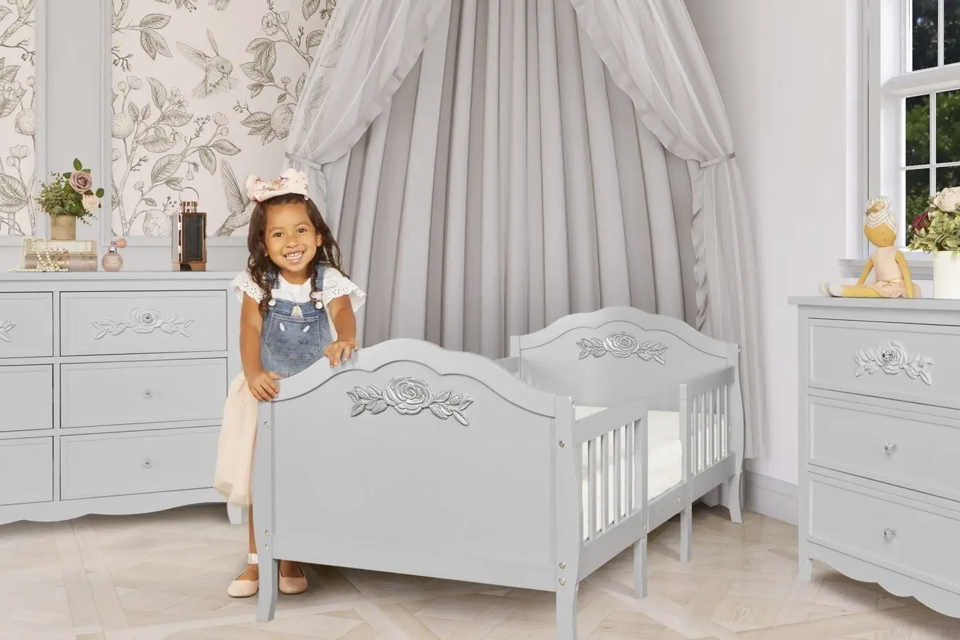 A young girl smiles while standing next to a gray toddler bed.