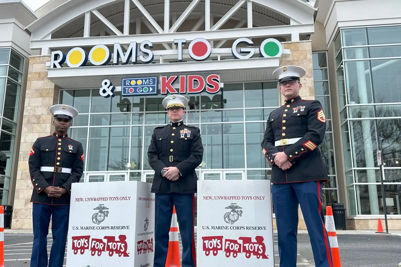 Three Marines in uniform standing in front of a Rooms To Go store with Toys for Tots boxes.
