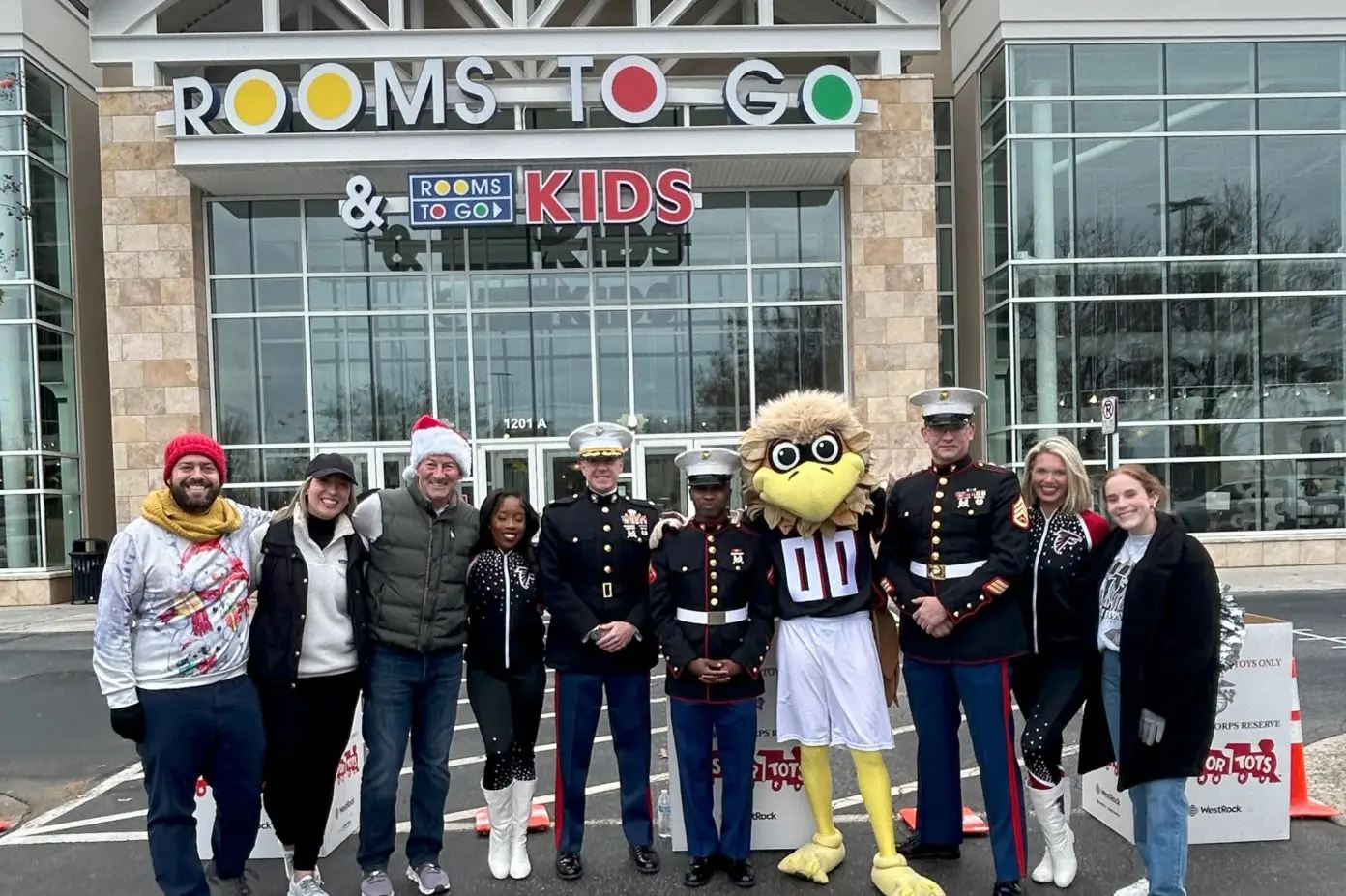 People standing in front of a Rooms To Go store with a mascot and military personnel.
