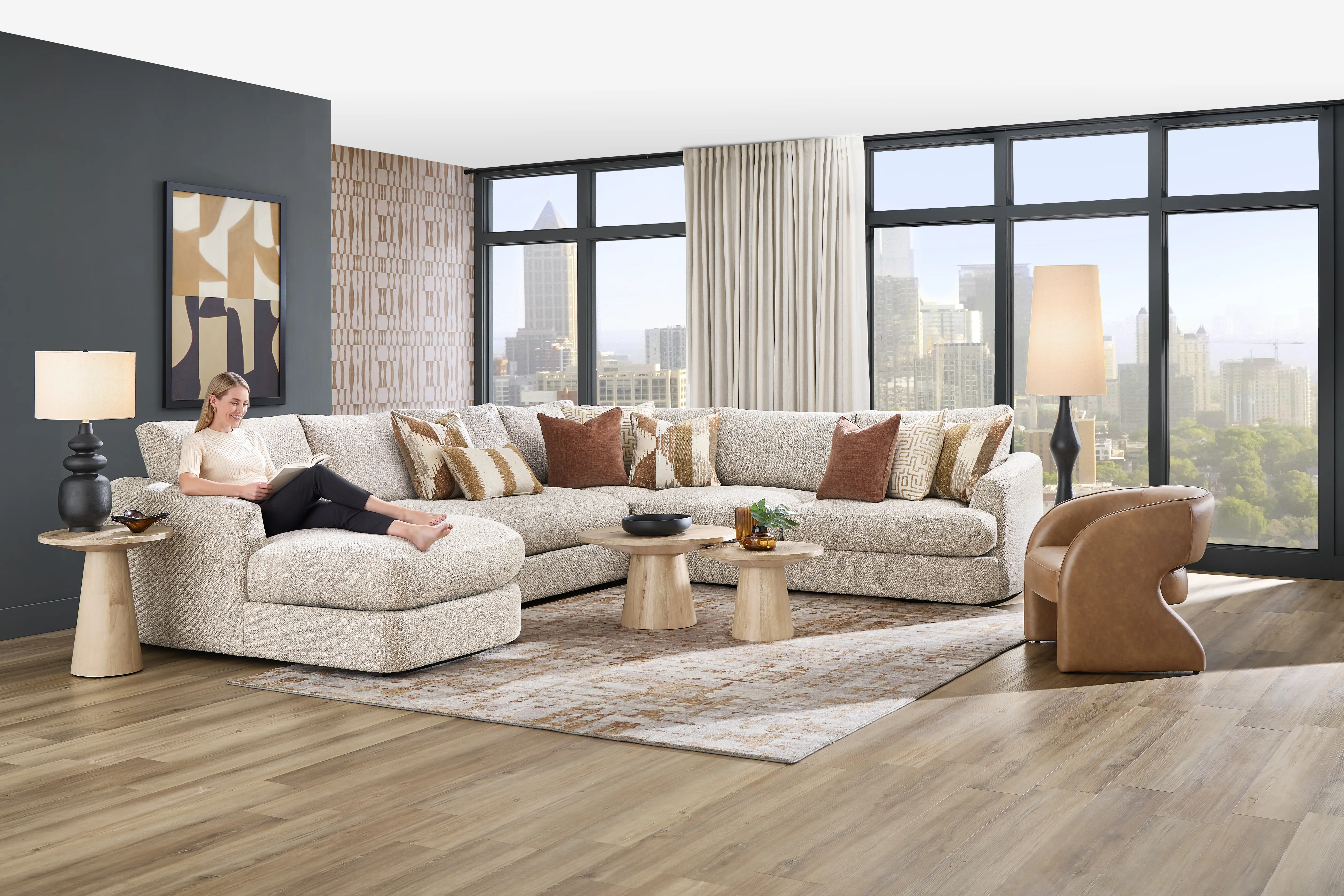 A woman sits on a large, beige sectional sofa in a modern living room with a city view, wooden coffee tables, and an armchair.