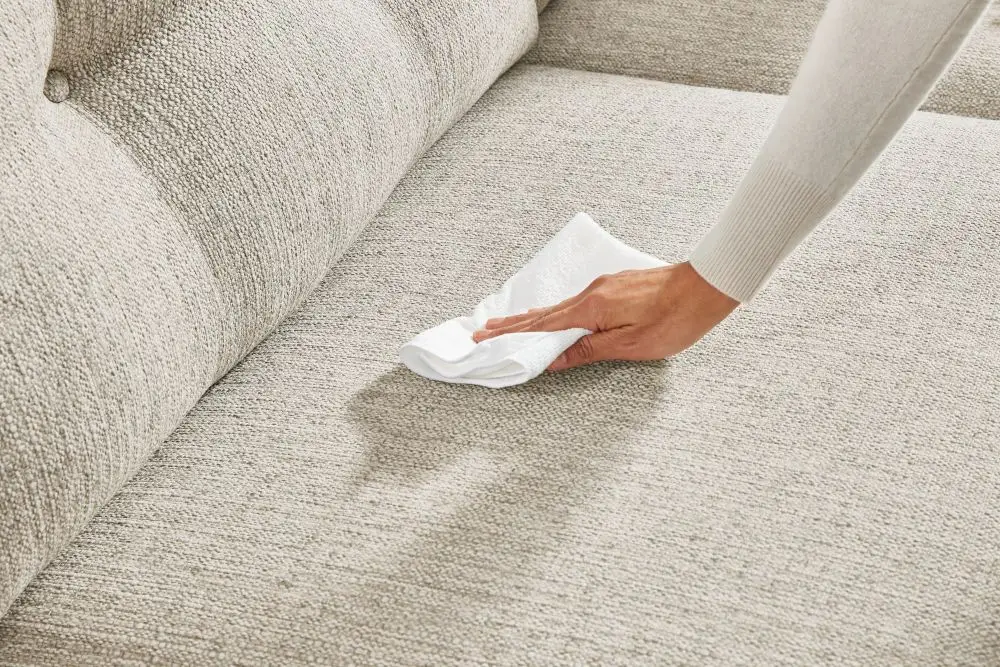 A hand uses a white cloth to clean a textured, light beige upholstered sofa.