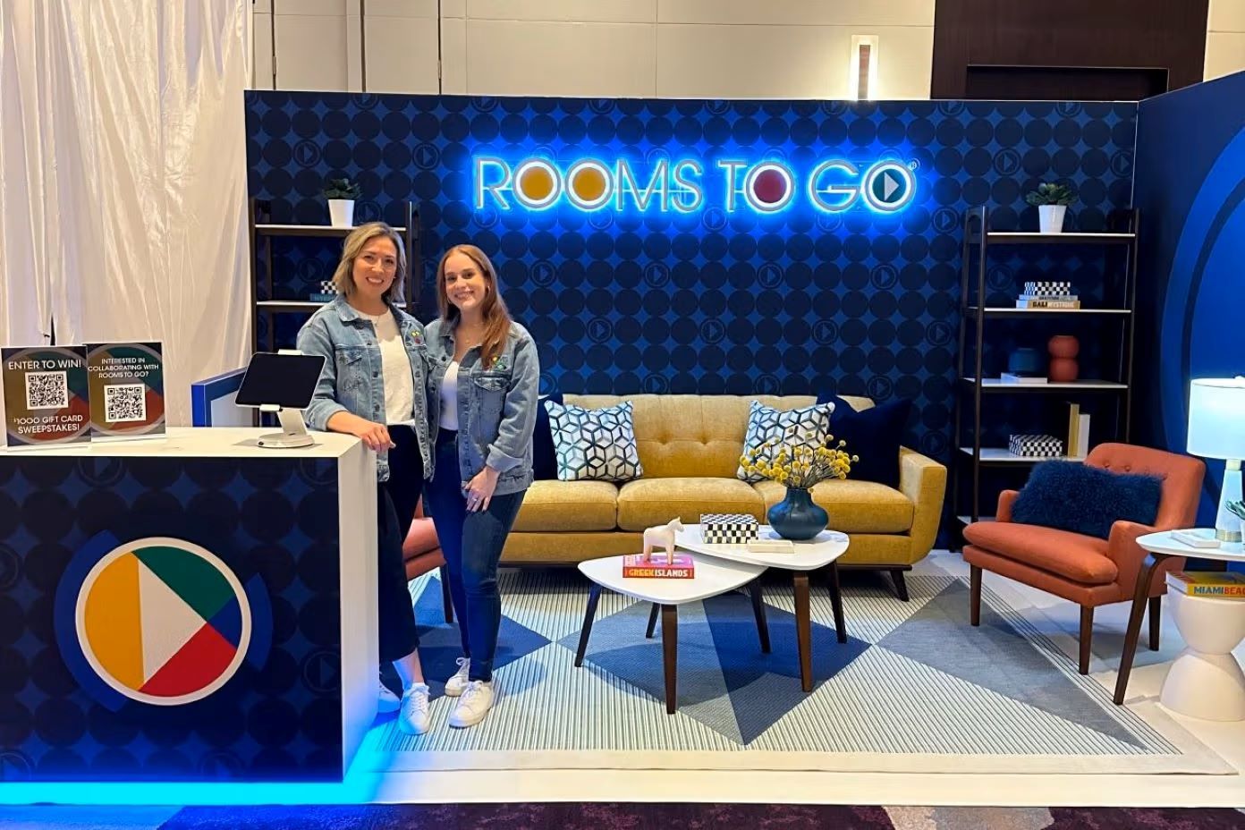 Two women standing in front of a Rooms To Go display.
