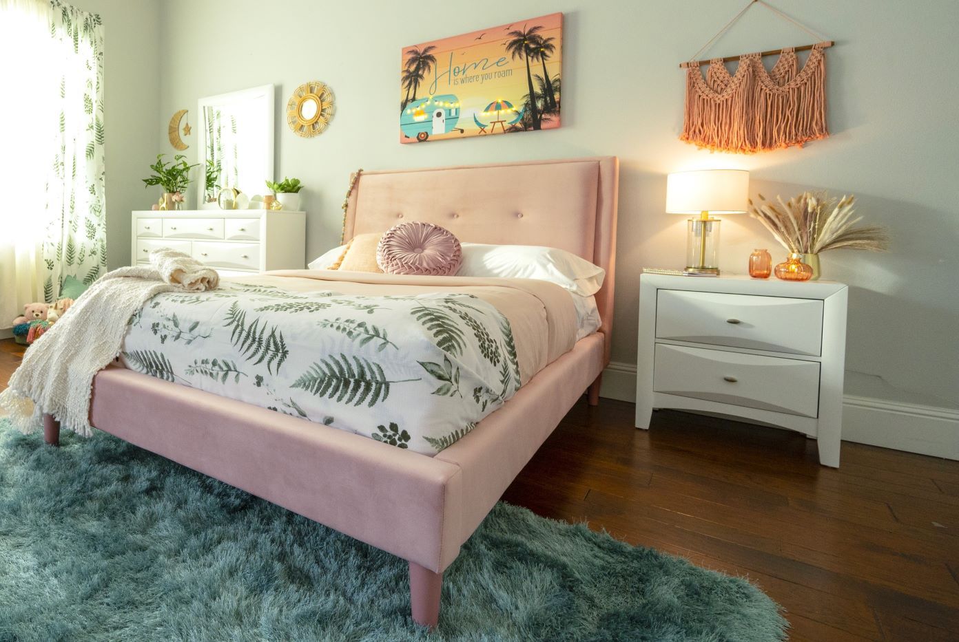 Pink bed in a bedroom with a blue rug and white dresser.
