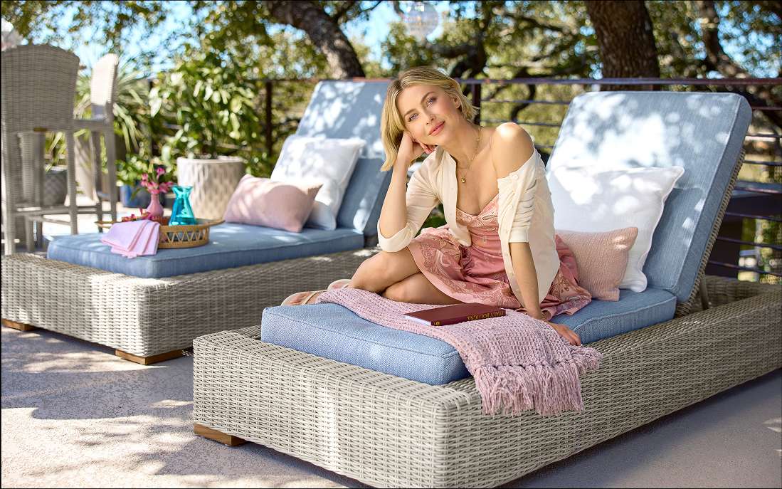 A woman sits on a wicker chaise lounge on a patio.
