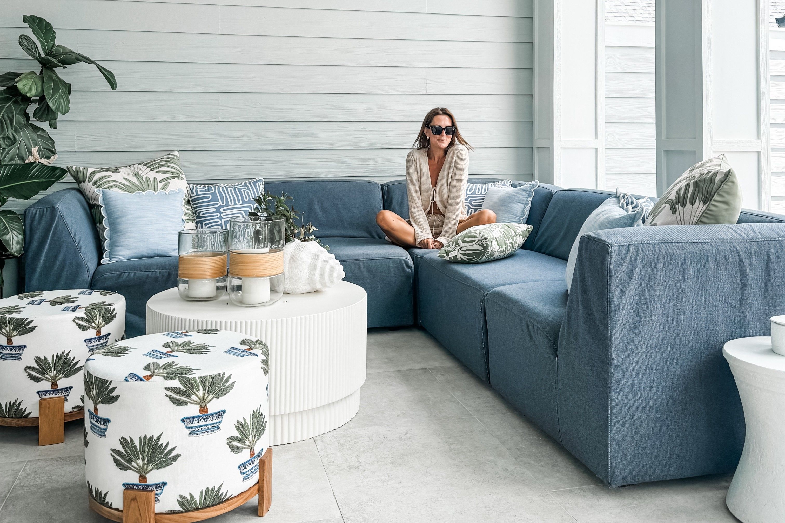 A woman sits on a blue sectional sofa on a patio. There are decorative pillows and two patterned ottomans.
