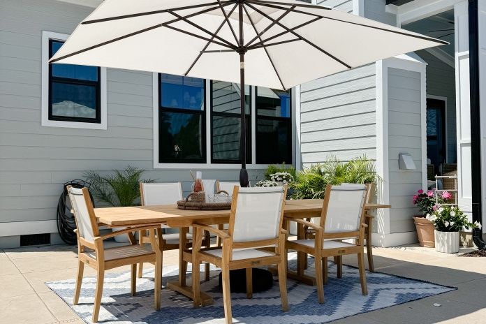 Modern patio dining setup with a wooden table, white chairs, and a large umbrella in a sunny backyard.