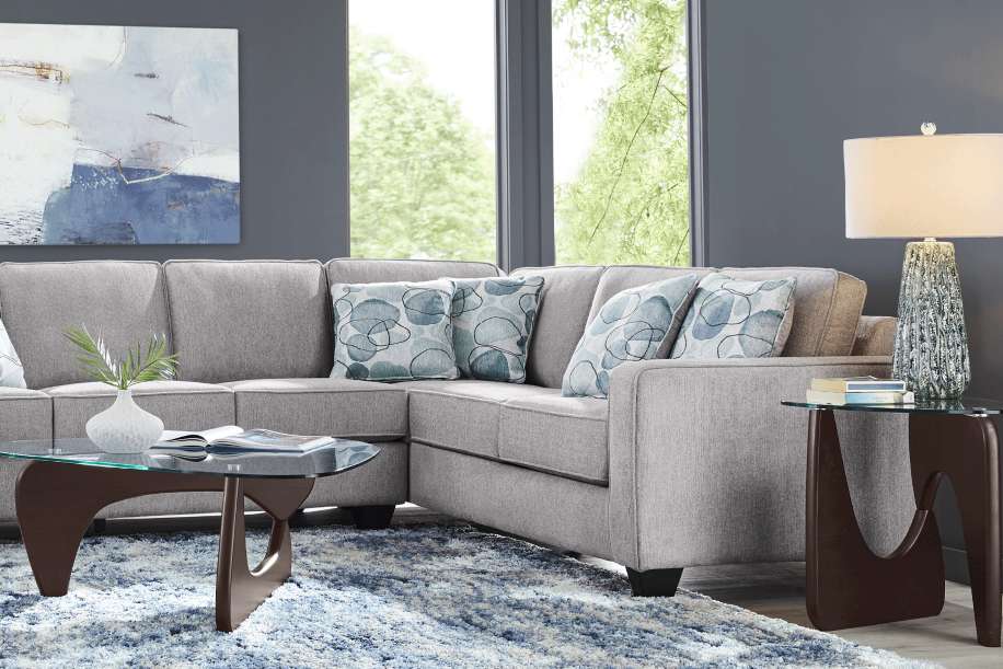 Gray sectional sofa with blue patterned pillows, a glass coffee table, and a blue rug.
