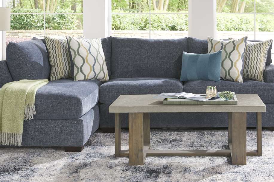Blue sectional sofa with patterned pillows and a green throw blanket. A gray coffee table sits in front.
