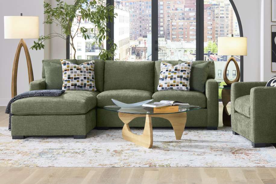 Green sectional sofa with patterned pillows, a glass coffee table, and a wooden floor lamp.