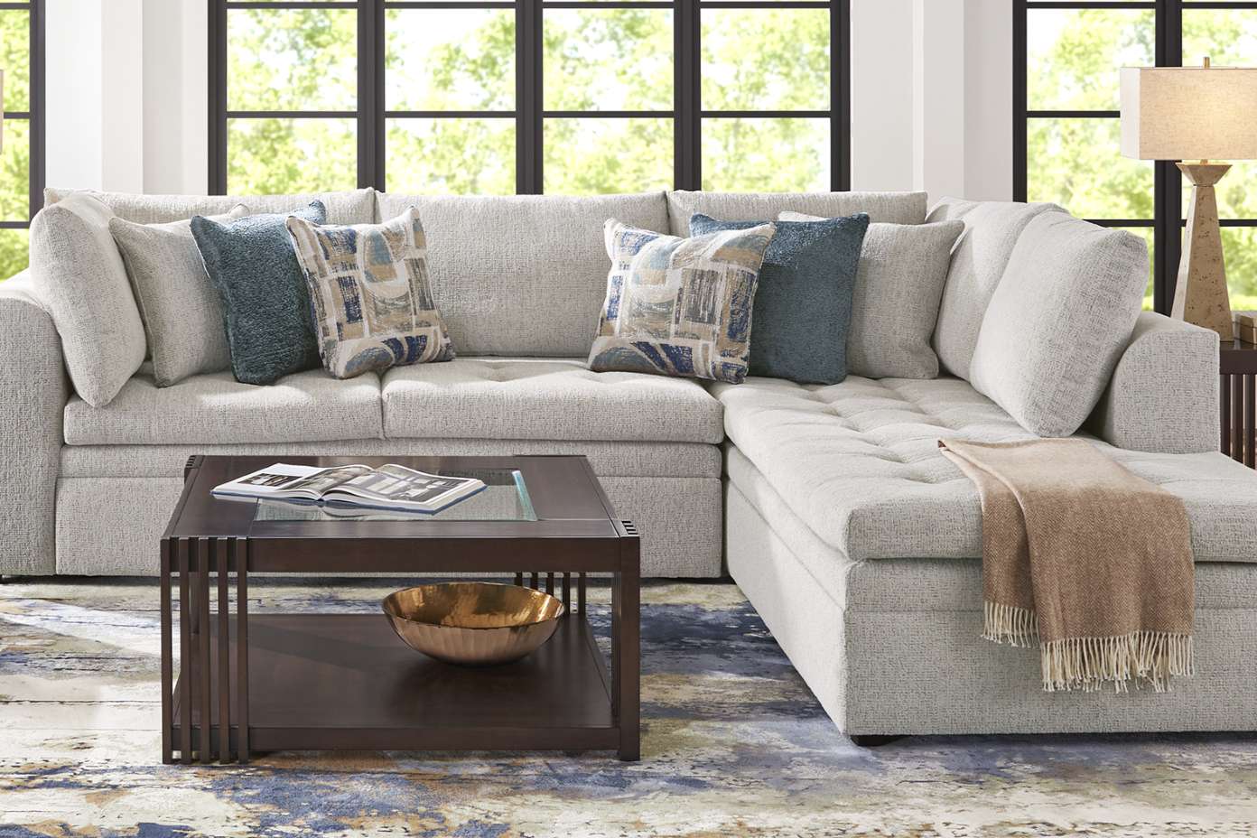 A light gray sectional sofa with patterned throw pillows and a brown throw blanket.
