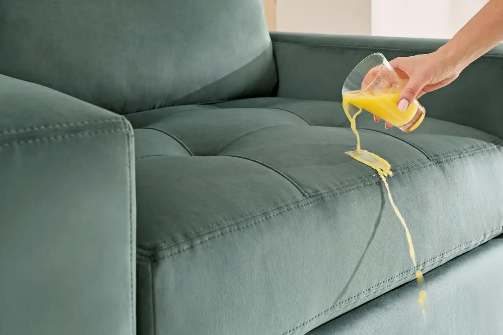 A hand pours orange juice onto a teal velvet sofa, demonstrating the fabric's liquid-repellent properties.