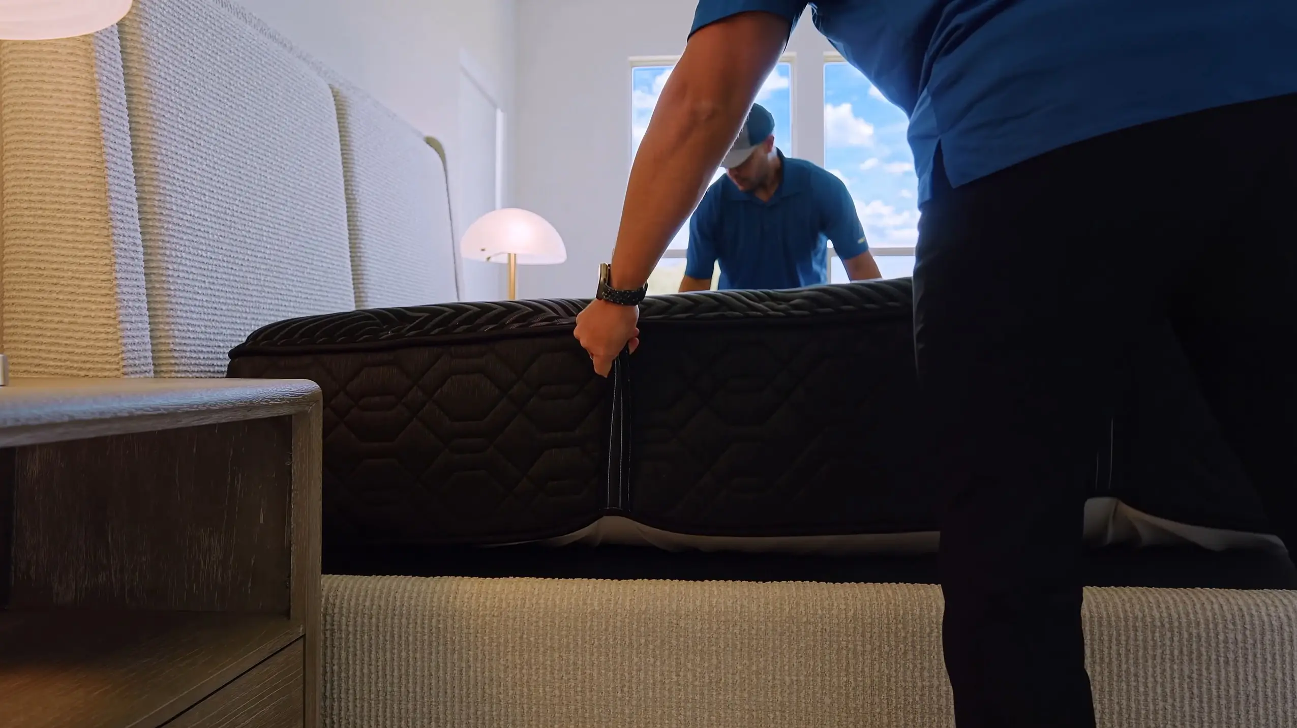 Two workers in blue uniforms position a dark, quilted mattress onto a textured, light-colored bed frame in a bright room.