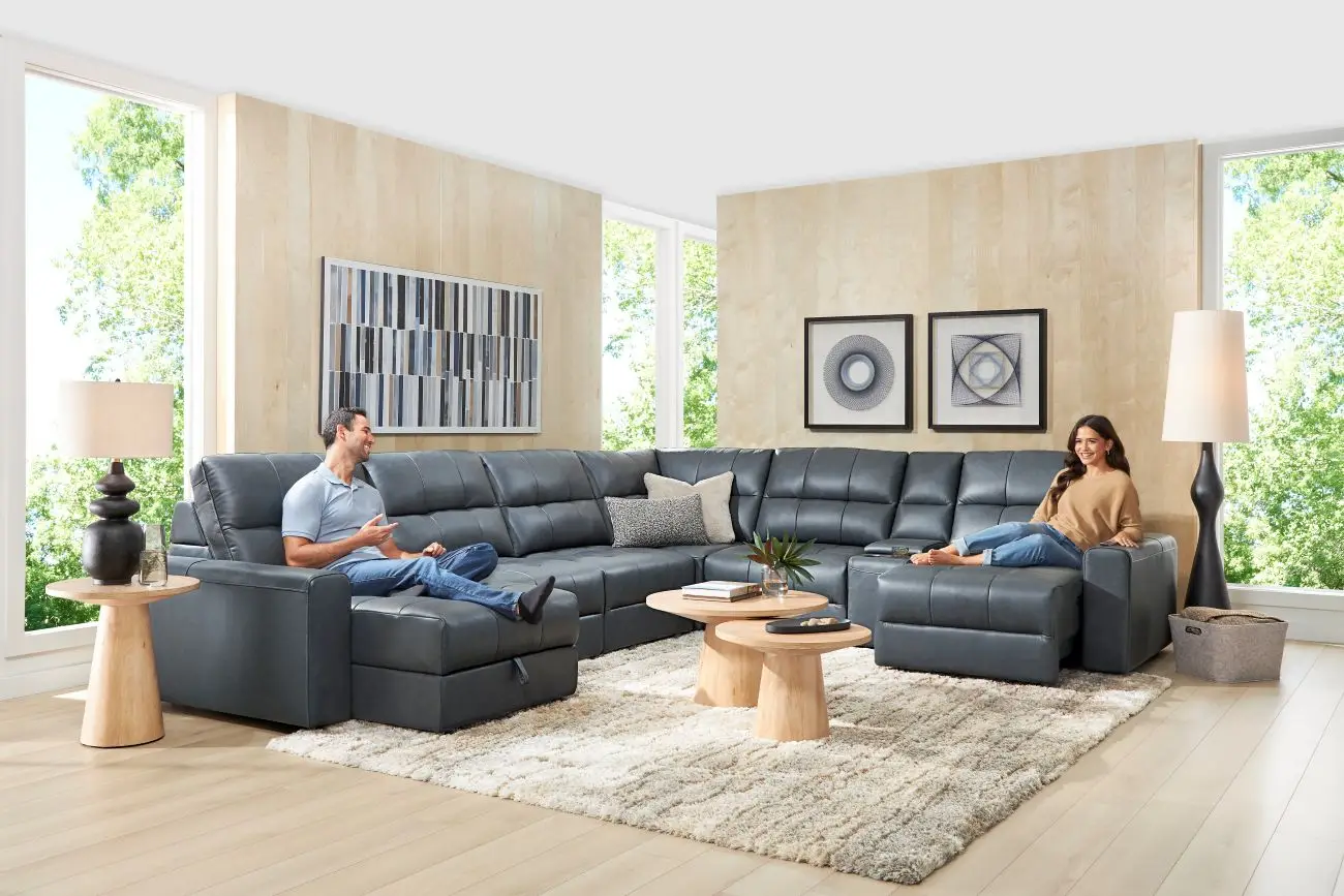 A man and woman sit on a large, dark gray sectional sofa in a bright, modern living room with wood-paneled walls.