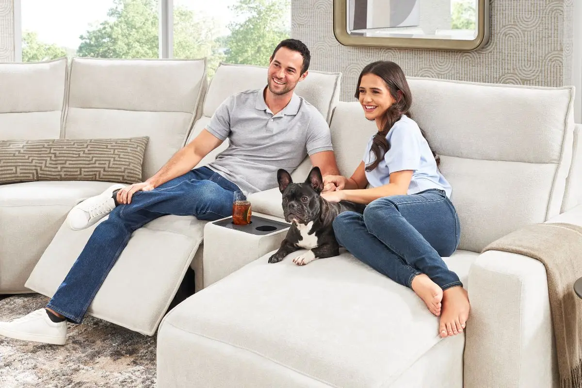 A man and woman sitting on a light-colored sectional sofa with a French bulldog, featuring a built-in cup holder.