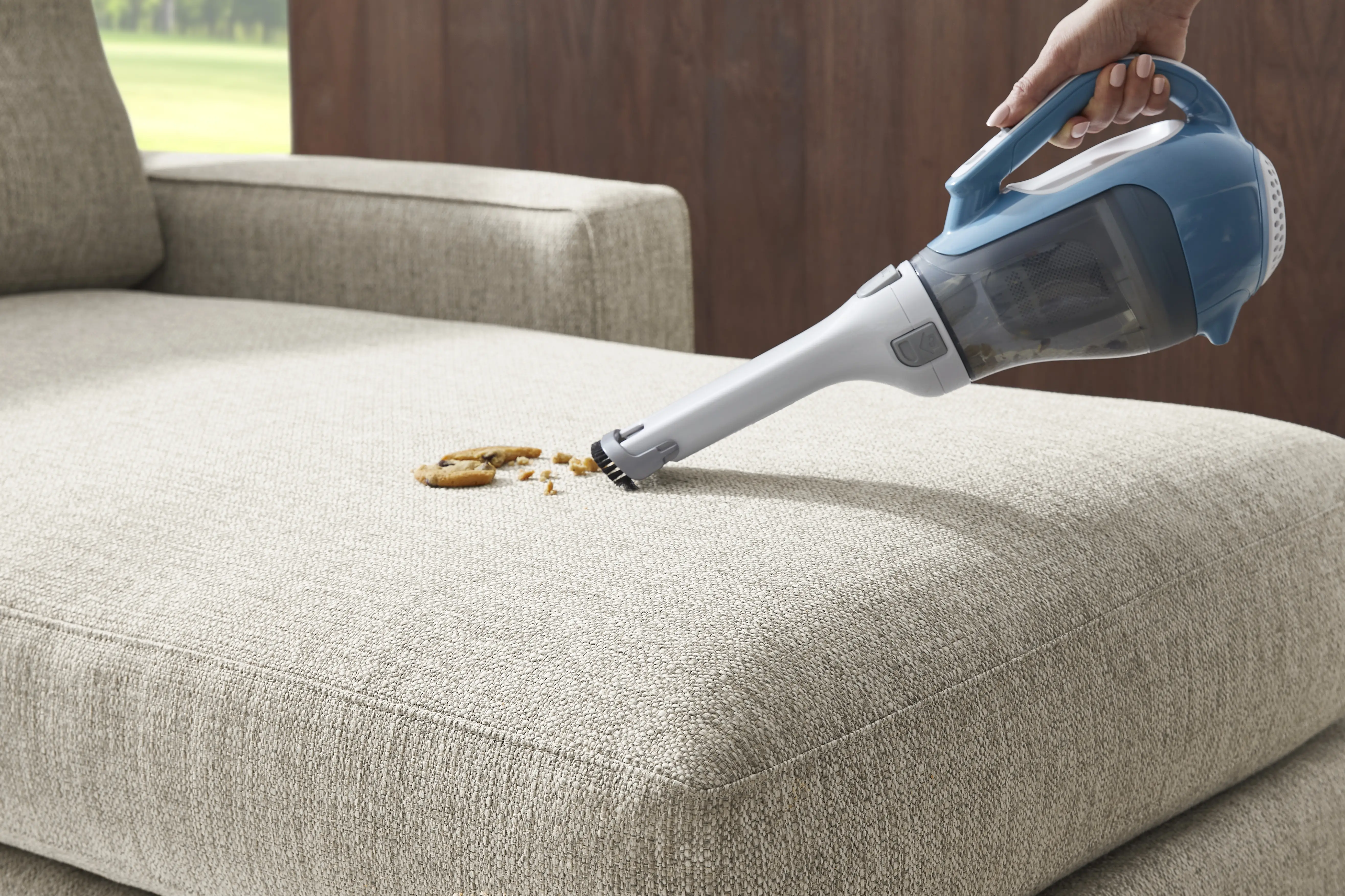 A hand uses a blue handheld vacuum to clean cookie crumbs off a beige fabric sofa.