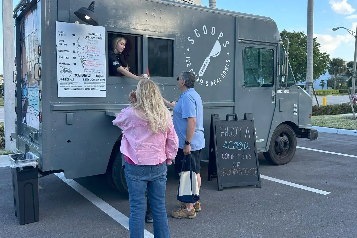 An ice cream truck with customers. The truck says "Scoops Ice Cream & Acai Bowls". A sign says "Enjoy a scoop compliments of Rooms to Go".
