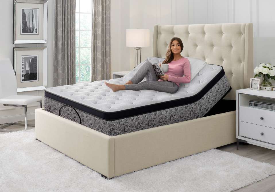 A woman sits on an adjustable bed, reading a magazine and holding a mug.
