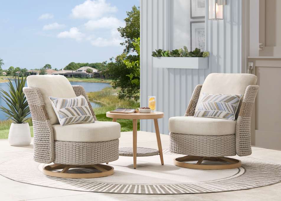 Two beige swivel chairs with pillows, a small table, and a rug on a patio.
