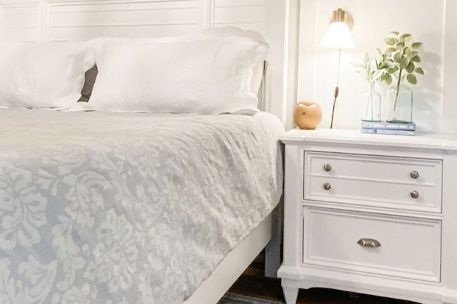 A white bedroom with a bed, nightstand, and lamp. The bed has white pillows and a patterned comforter.
