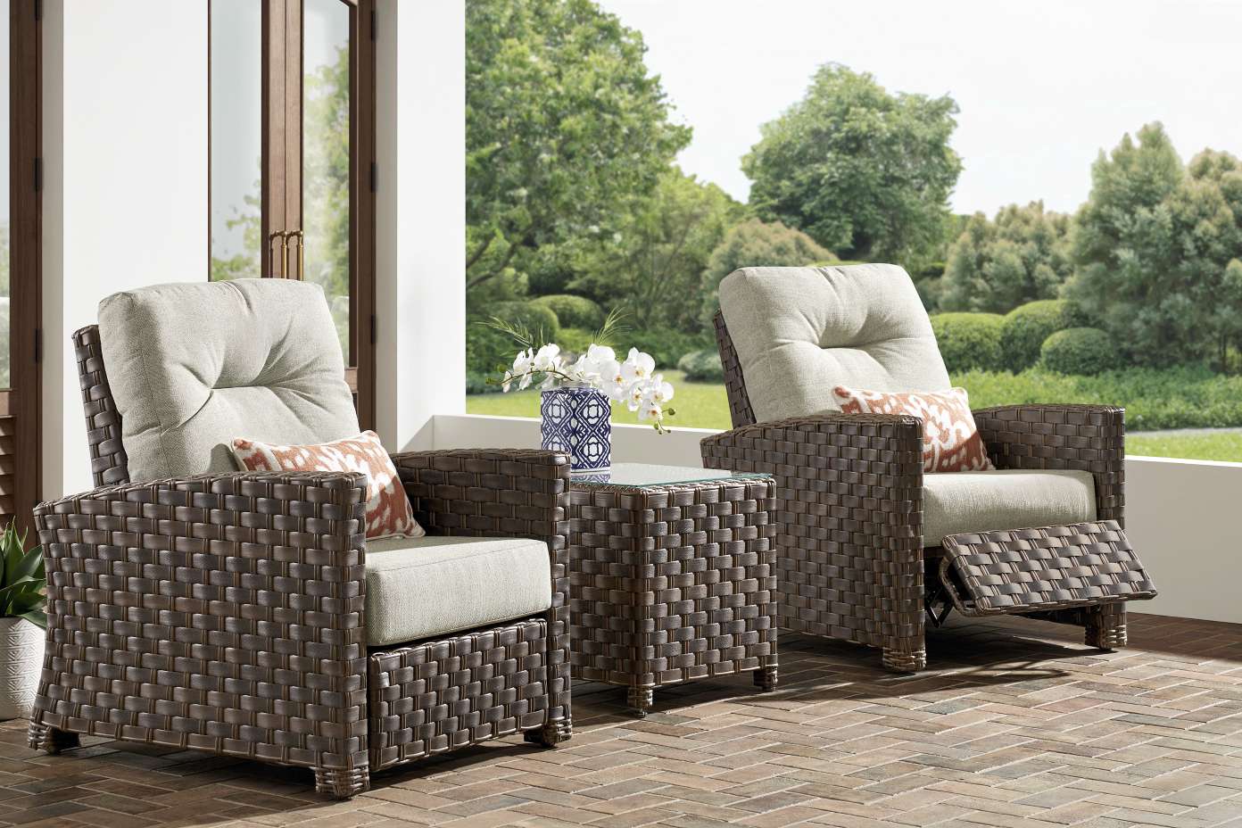 Two brown wicker outdoor recliner chairs with light green cushions and patterned pillows, with a small side table.
