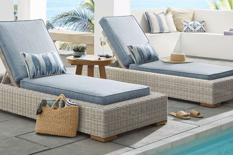 Two outdoor chaise lounges with blue cushions and pillows, next to a pool.
