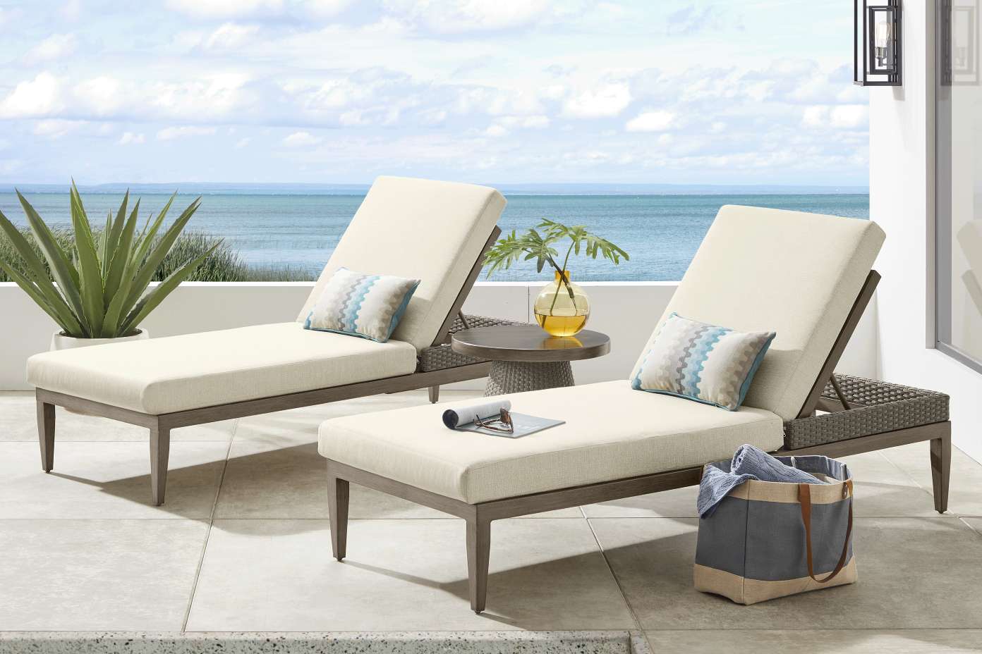 Two beige chaise lounges with pillows, a side table, and a tote bag on a patio overlooking the ocean.
