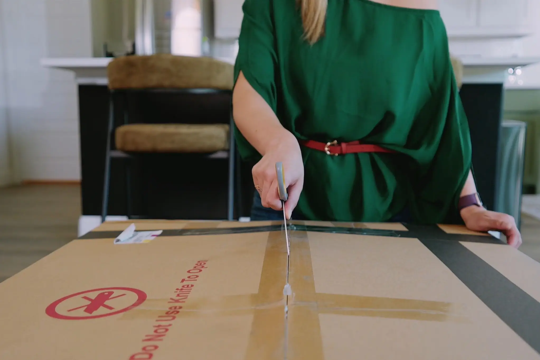 Do Not Use Knife To Open. A person in a green shirt uses scissors to cut open a cardboard box.