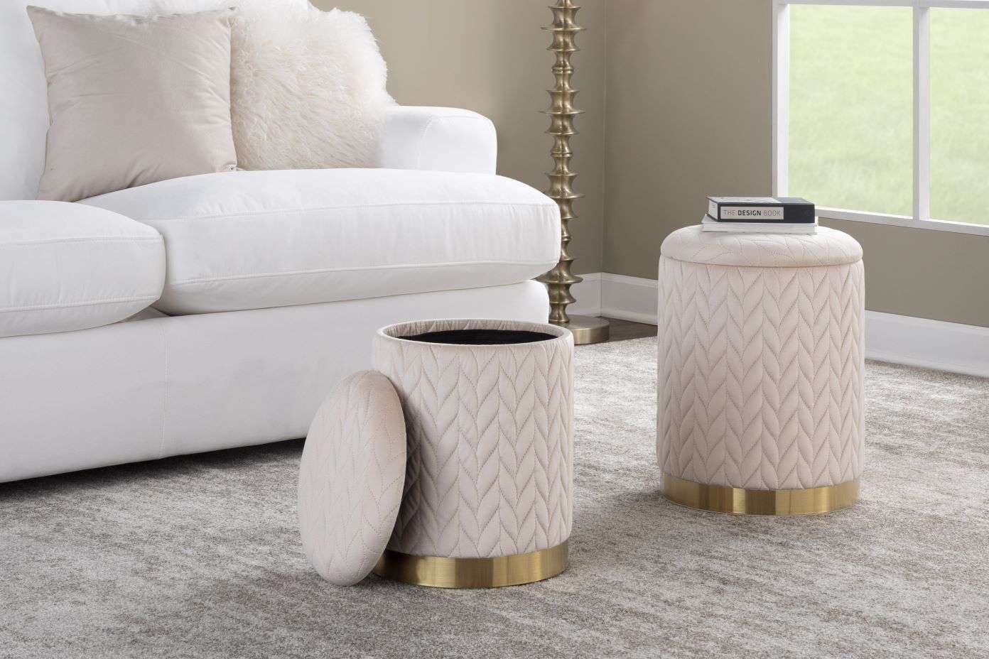 Two cream-colored storage ottomans with gold bases, one with its lid open, next to a white sofa.