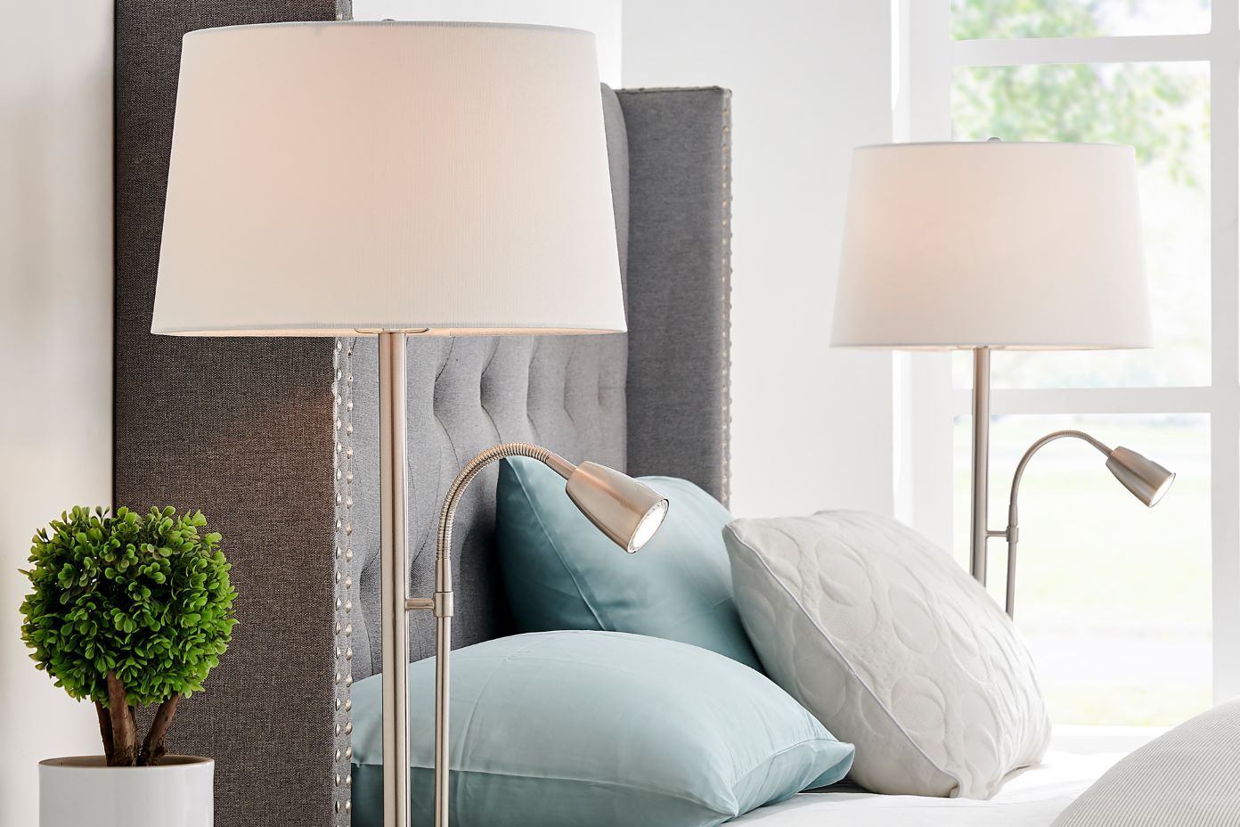 Floor lamps with white shades and flexible reading lights next to a bed with pillows.

