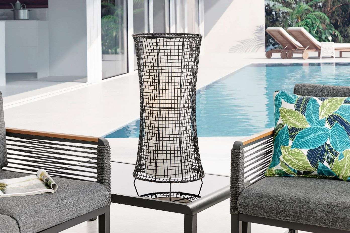 Black wire lantern on a table between two gray outdoor chairs with a pool in the background.
