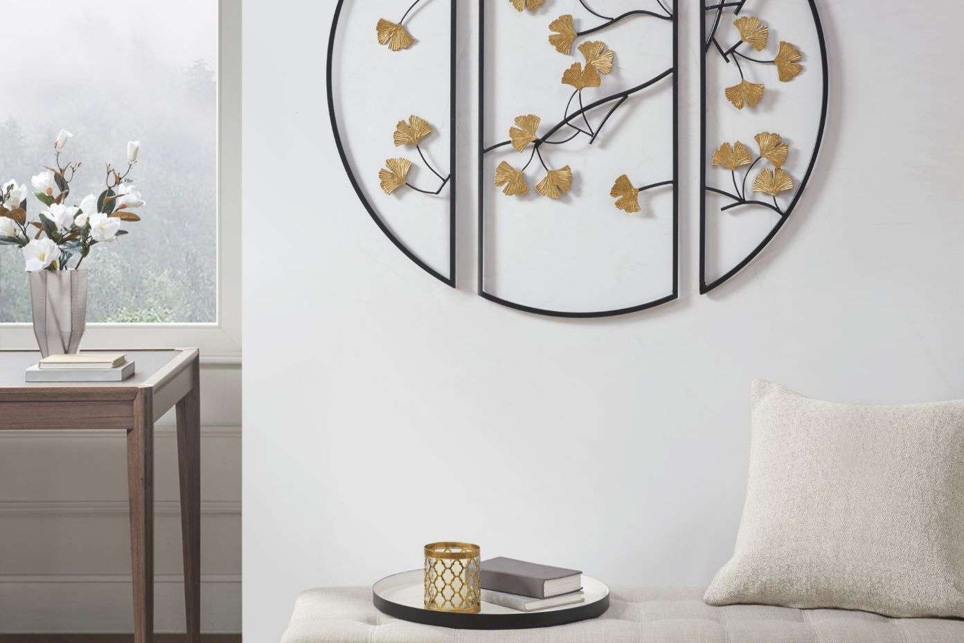 A wall decoration with gold leaves, a table with a vase of flowers, and a bench with a pillow.
