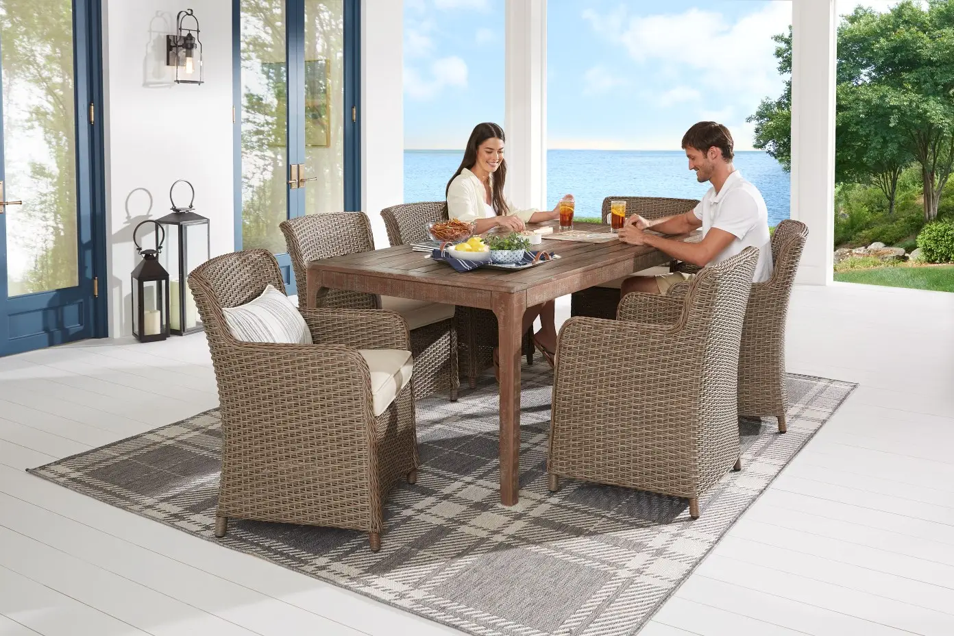 A couple sits at a rustic wooden outdoor dining table with wicker chairs on a plaid rug on a patio overlooking the sea.