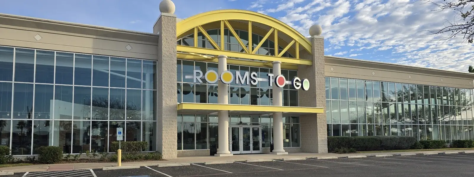 Rooms To Go storefront with a large glass facade and a yellow arched entrance structure under a blue sky.