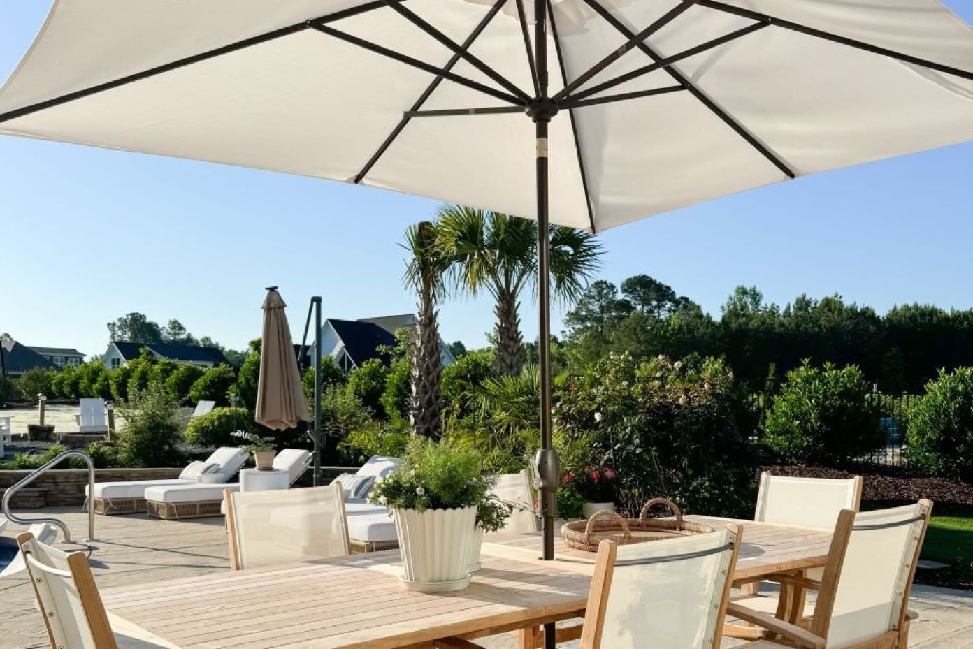 A patio table and chairs are under a large umbrella. In the background are palm trees and a pool.
