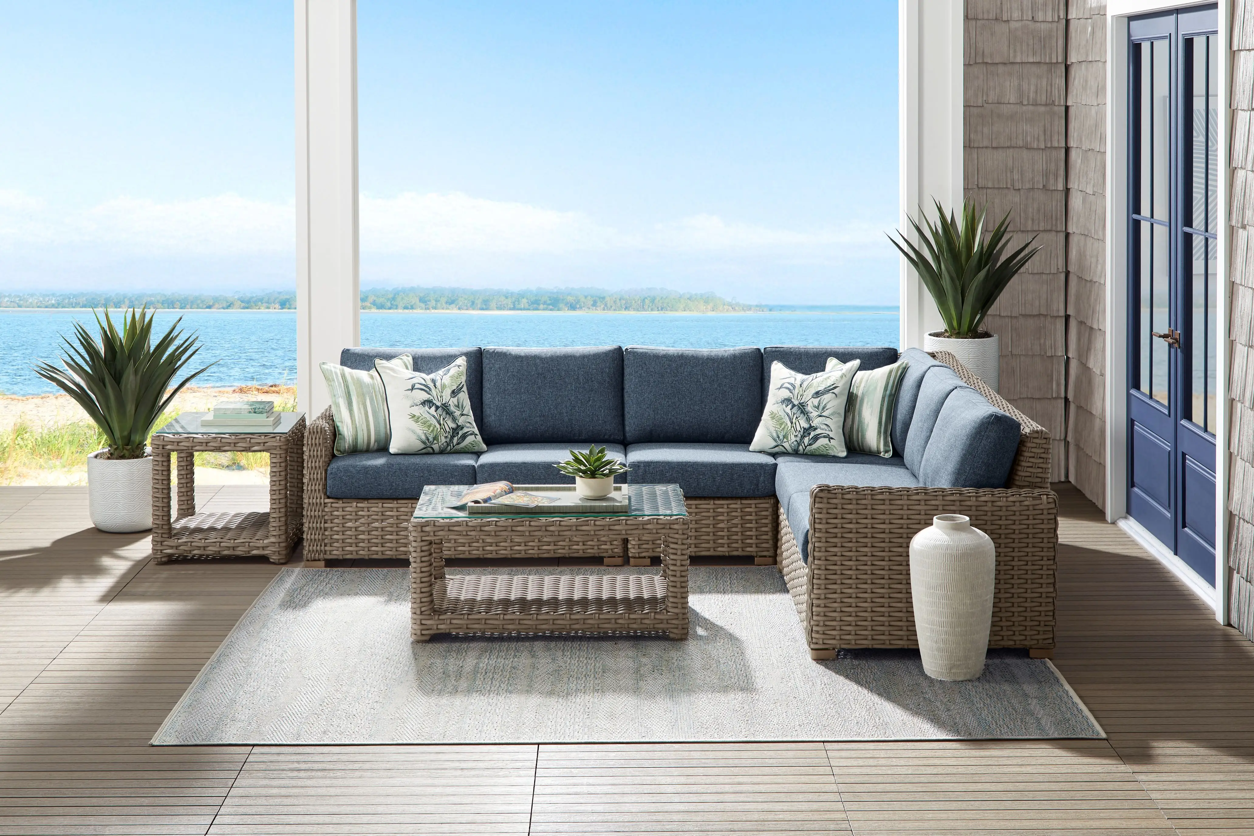 A blue sectional sofa with patterned pillows and a wicker coffee table on a patio overlooking the ocean.
