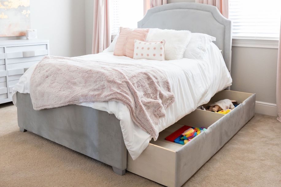 A gray upholstered bed with storage drawers, white bedding, and pink pillows.
