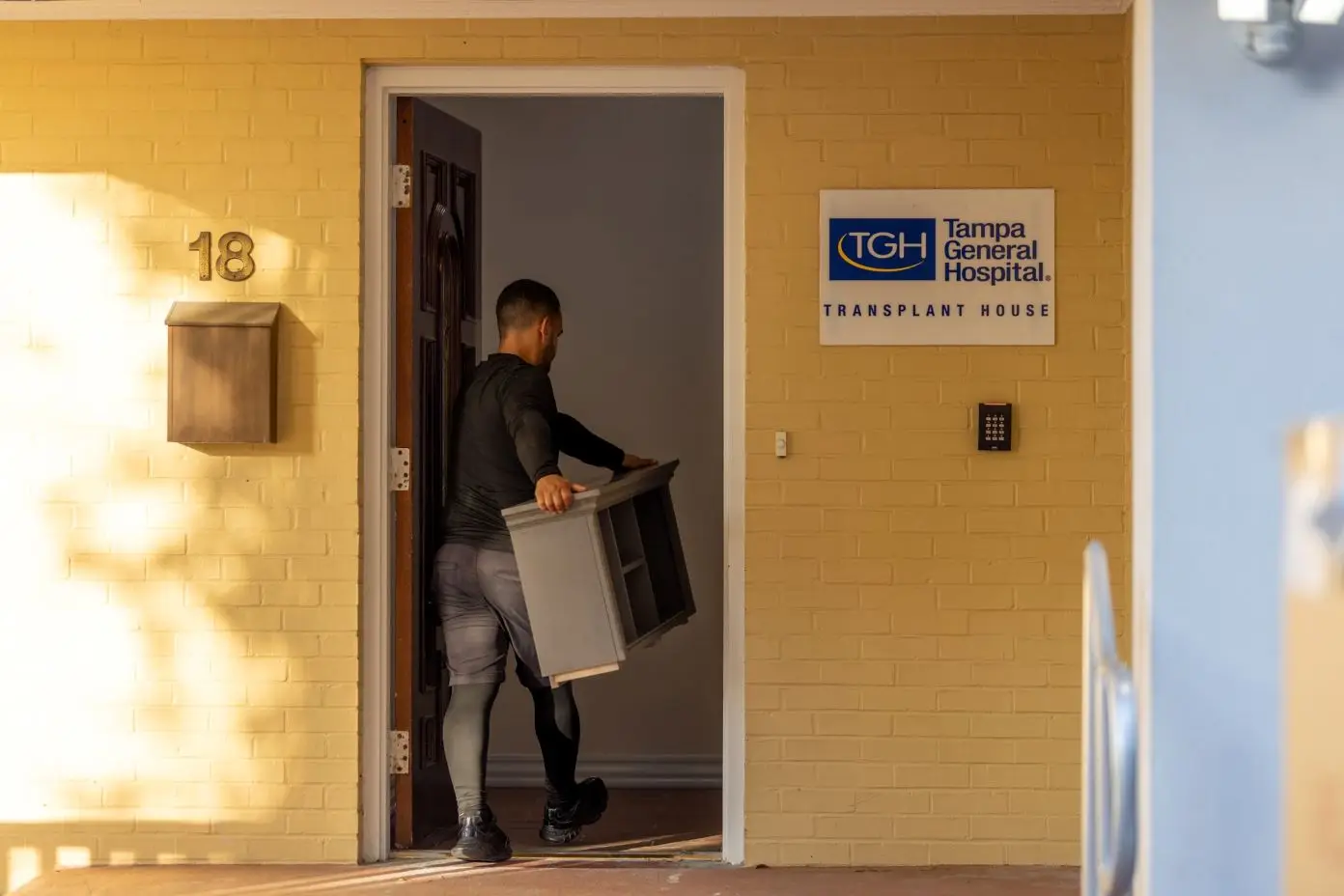 Tampa General Hospital Transplant House sign. A man carries a gray cabinet through the doorway.
