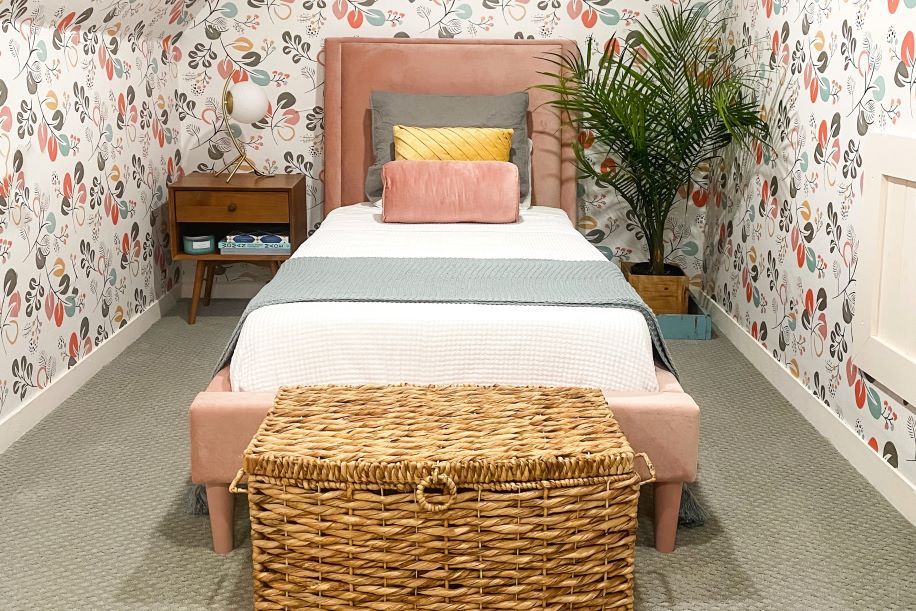 A bedroom with a pink bed, floral wallpaper, and a woven basket.
